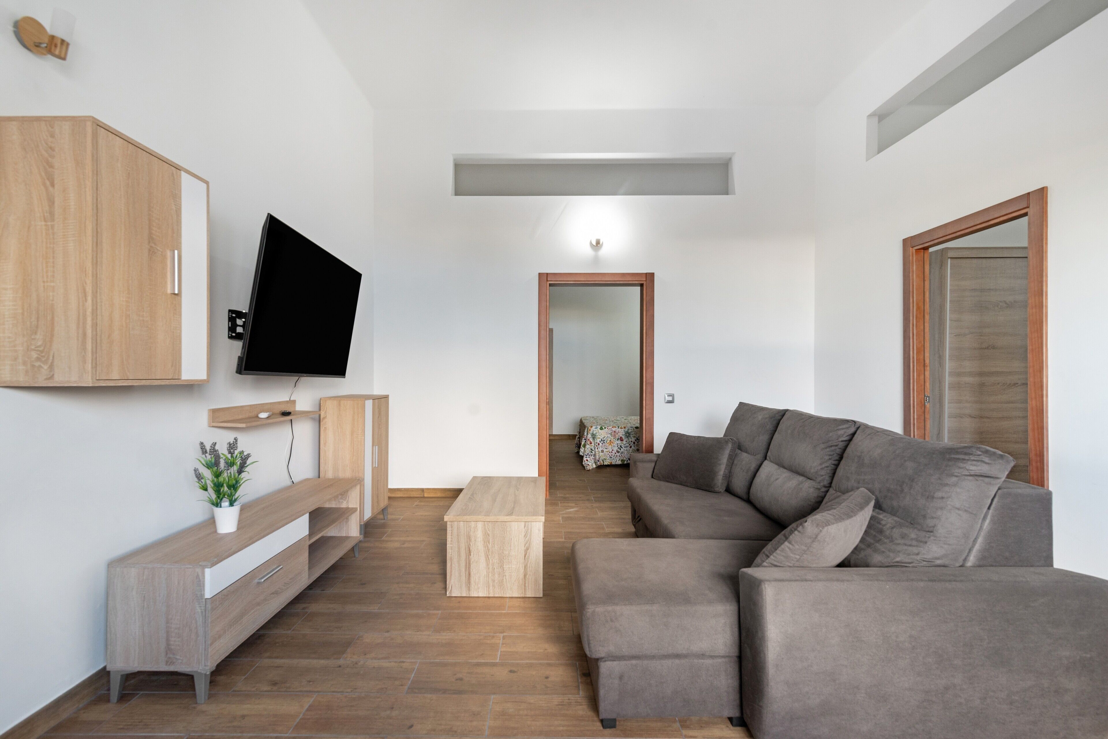 Een moderne woonkamer met een grijze bank, een houten salontafel, een aan de muur bevestigde televisie en een houten kast.