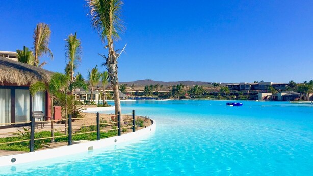A large, clear blue swimming pool surrounded by palm trees and resort buildings.