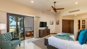 A bedroom with a bed, a sofa, a TV, and a sliding glass door leading to a balcony with a view of the beach.