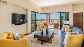 A modern living room with a flat-screen TV, two sofas, a coffee table, and a large sliding glass door leading to a balcony with a view of the sea.