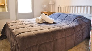 A neatly made bed with a brown duvet and a rolled white towel on top, a bedside table with a lamp, and a window with curtains.