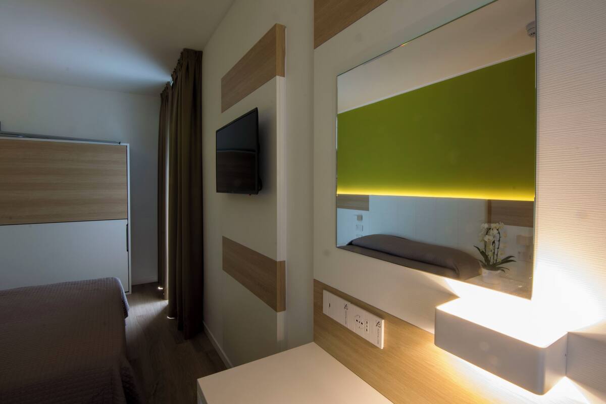 Een moderne hotelkamer met een bed, een aan de muur bevestigde televisie, een nachtkastje met spiegel en een kleine plant op het nachtkastje.