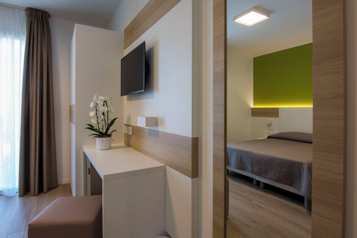 Een moderne hotelkamer met een flatscreen televisie aan de wand, een klein bureau met een bloeiende orchidee, een bed met een groene accentmuur, en een houten vloer.
