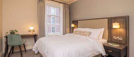 A bedroom with a large bed, a desk with a chair, a window with curtains, and a chandelier.