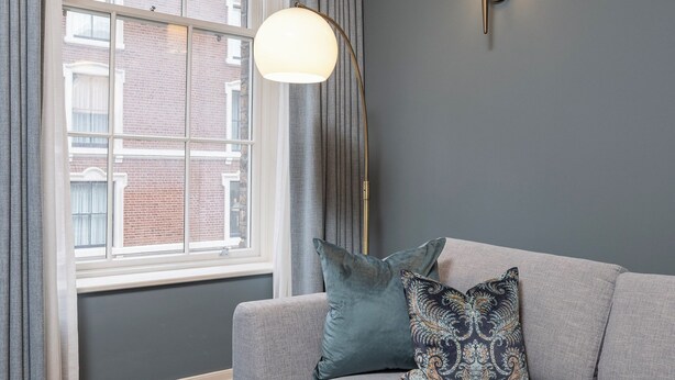 Un salon moderne avec un canapé d’angle, une lampe sur pied et une vue sur un bâtiment en briques, visible à travers une grande fenêtre.
