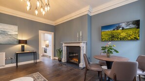 Un salon avec une cheminée, une table à manger avec des chaises, un grand tableau et un lustre.