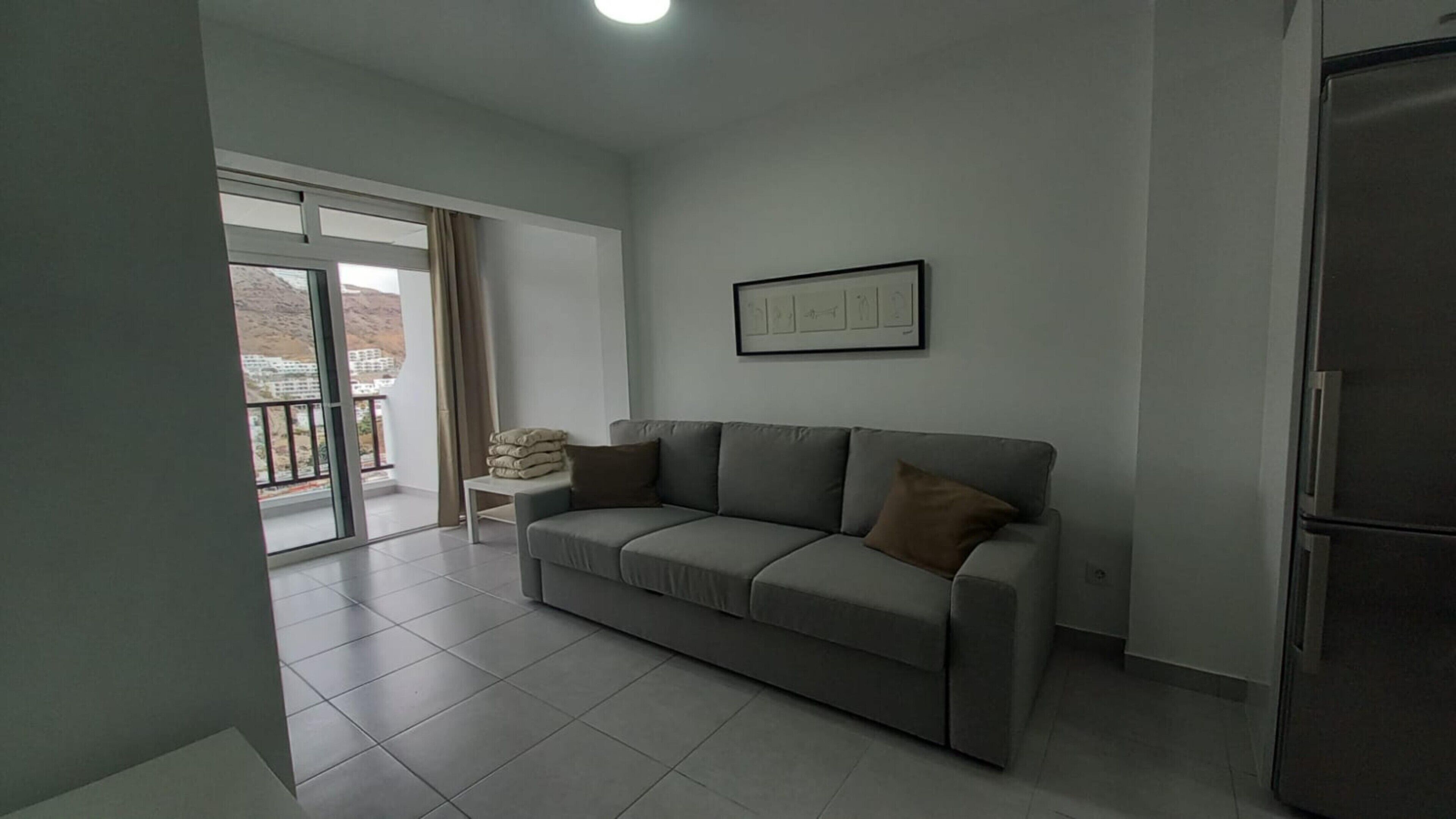 A living room with a grey sofa, a small table with cushions, a framed picture on the wall, and a sliding glass door leading to a balcony.