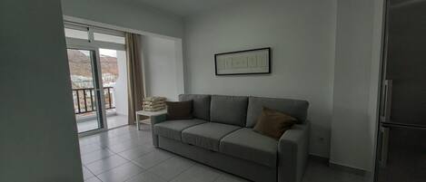 A living room with a grey sofa, a small table with cushions, a framed picture on the wall, and a sliding glass door leading to a balcony.