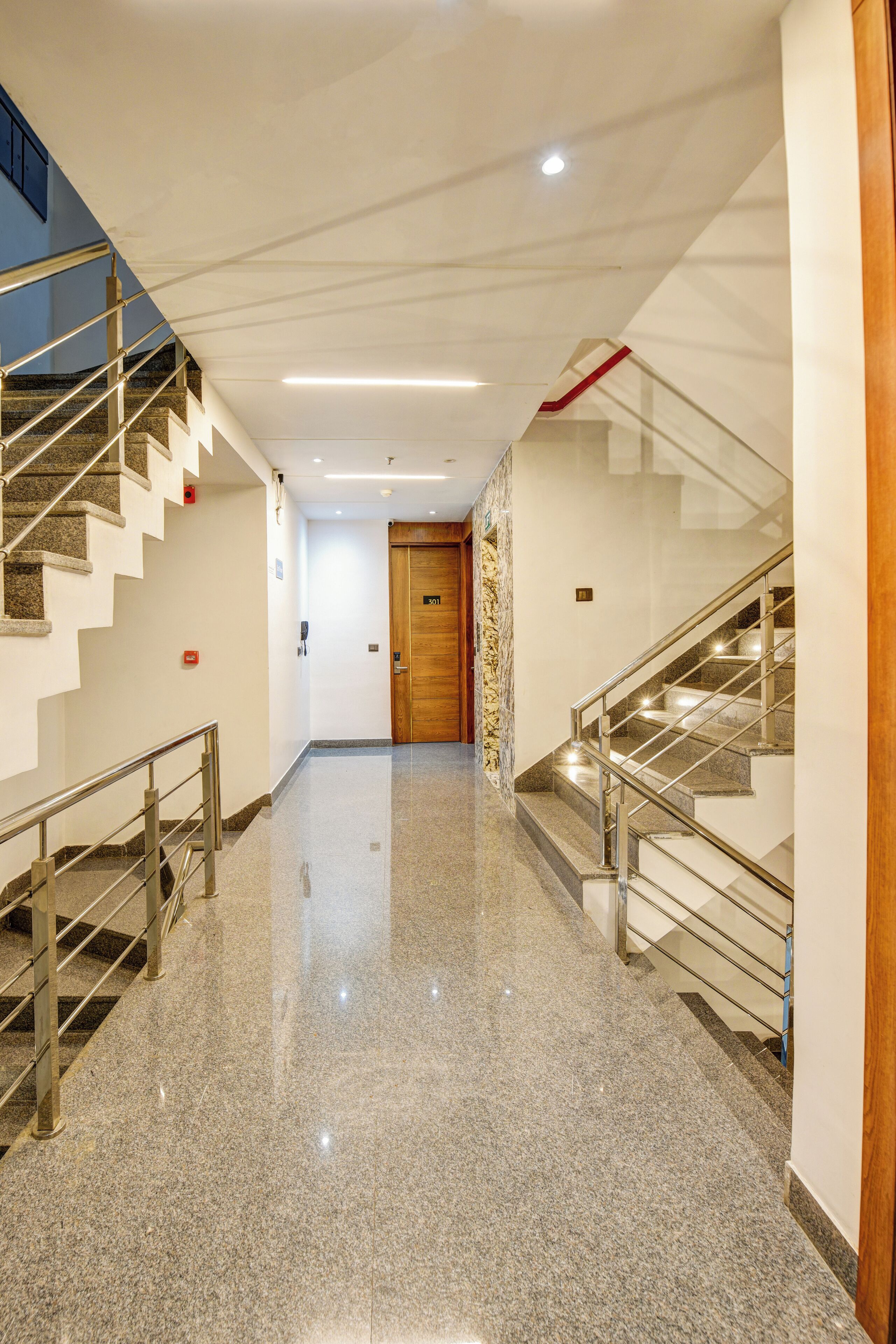 A clean, well-lit hallway with polished granite floors, a wooden door at the end, and two staircases with metal railings.