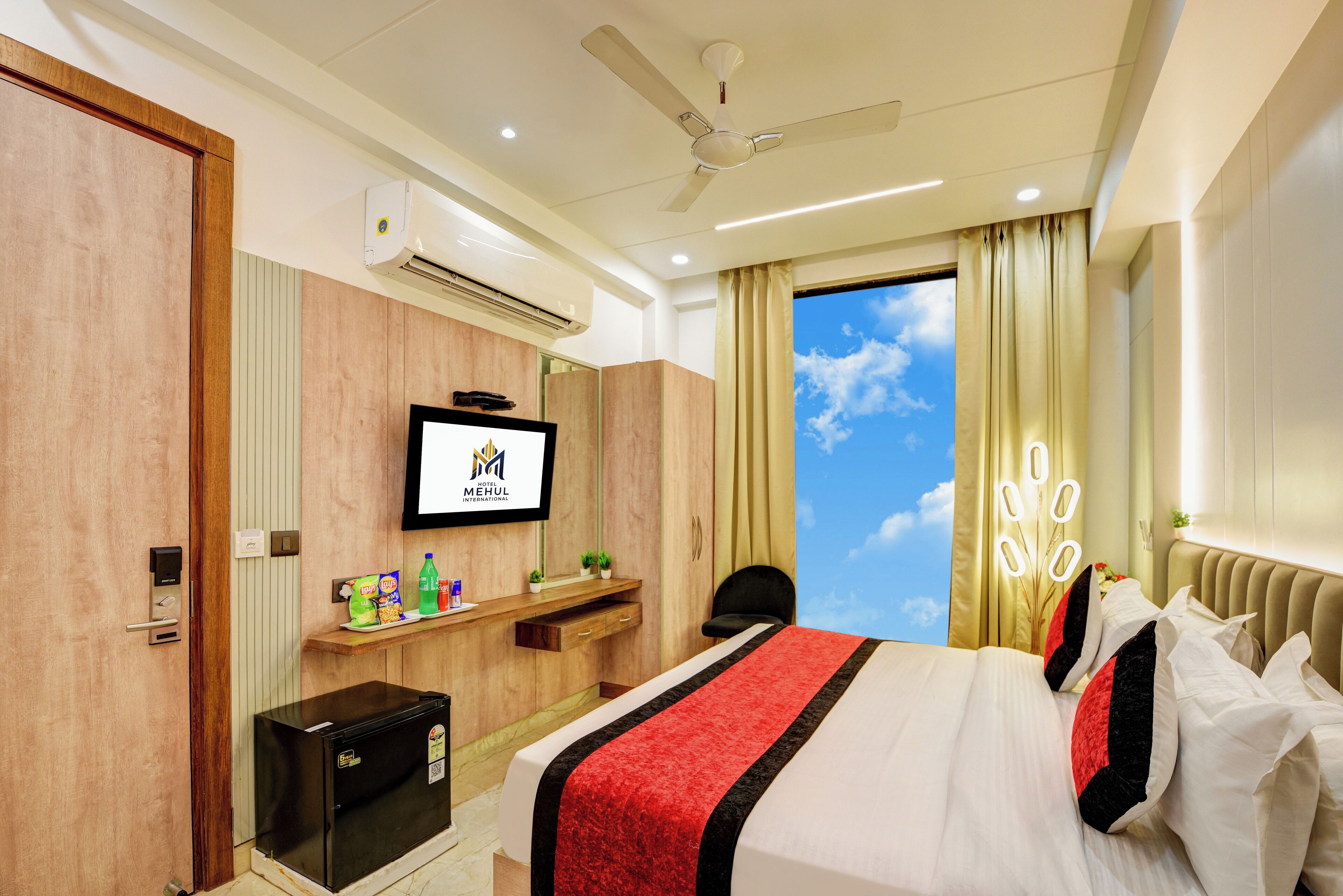 A hotel room with a bed, a TV, a minibar, a ceiling fan, and a window with curtains.