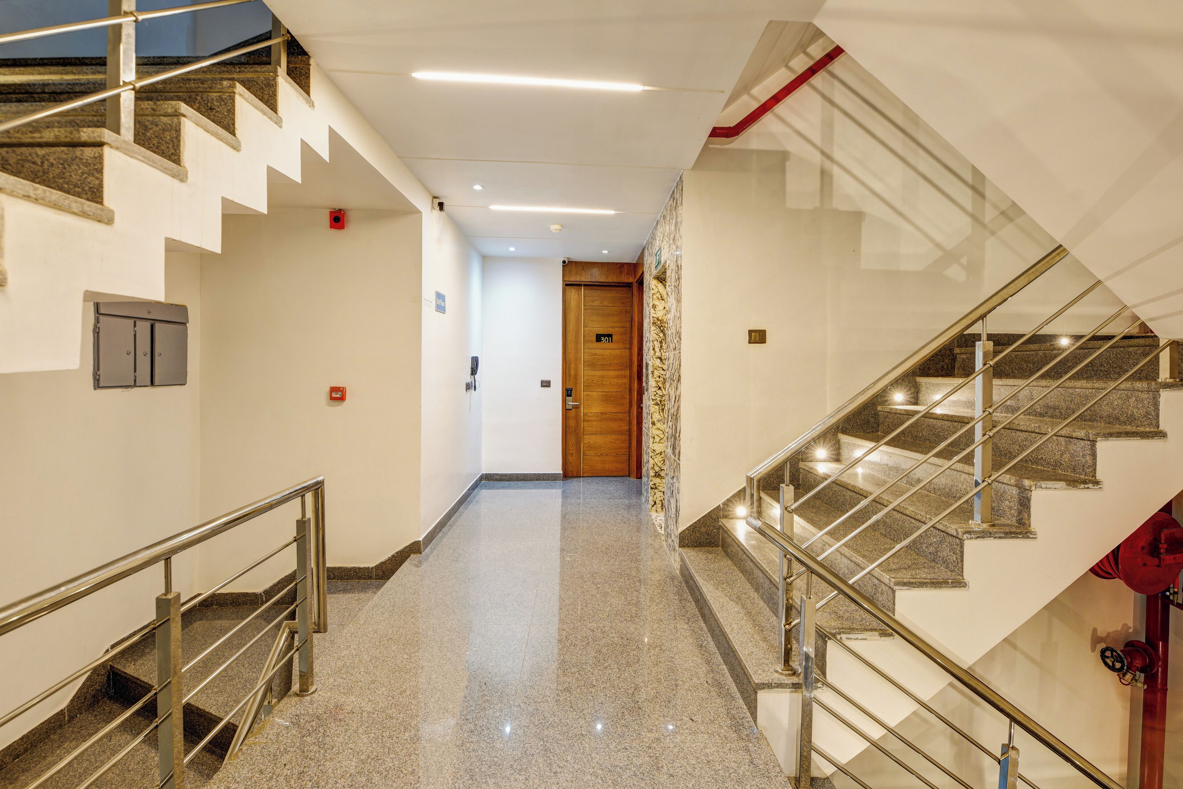 A hotel corridor with a polished granite floor, a wooden door at the end, and a staircase with metal railings.