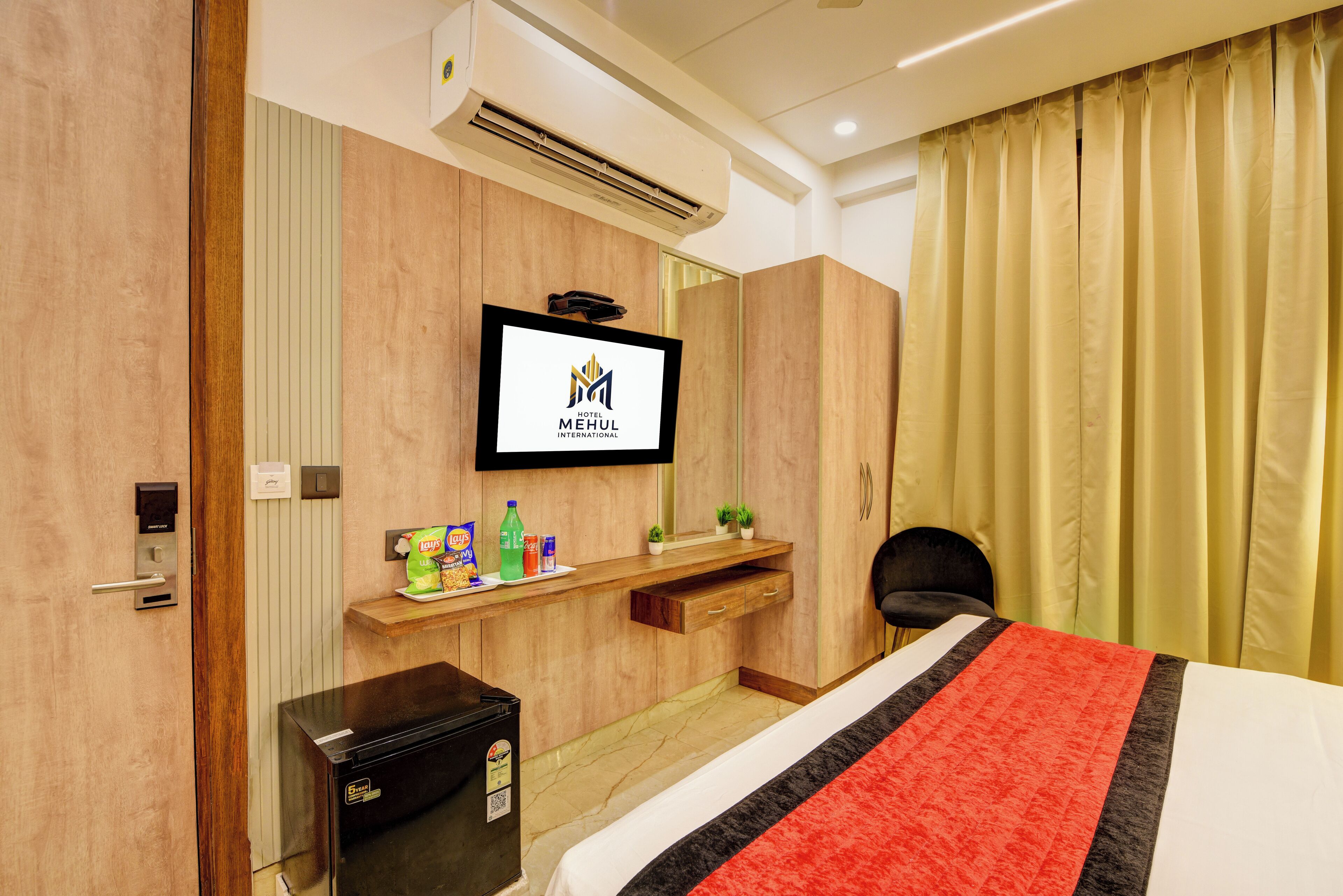 A hotel room with a bed, a TV mounted on the wall, a minibar, and a desk with a plant and snacks.