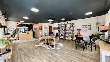 A cozy restaurant with wooden flooring, a central dining table with chairs, a bar area, and a decorative bookshelf.