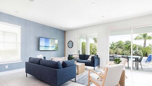 Una sala de estar moderna con un sofá azul, un sillón blanco y una mesa de centro. Hay un televisor montado en la pared y un ventanal con vistas al exterior.