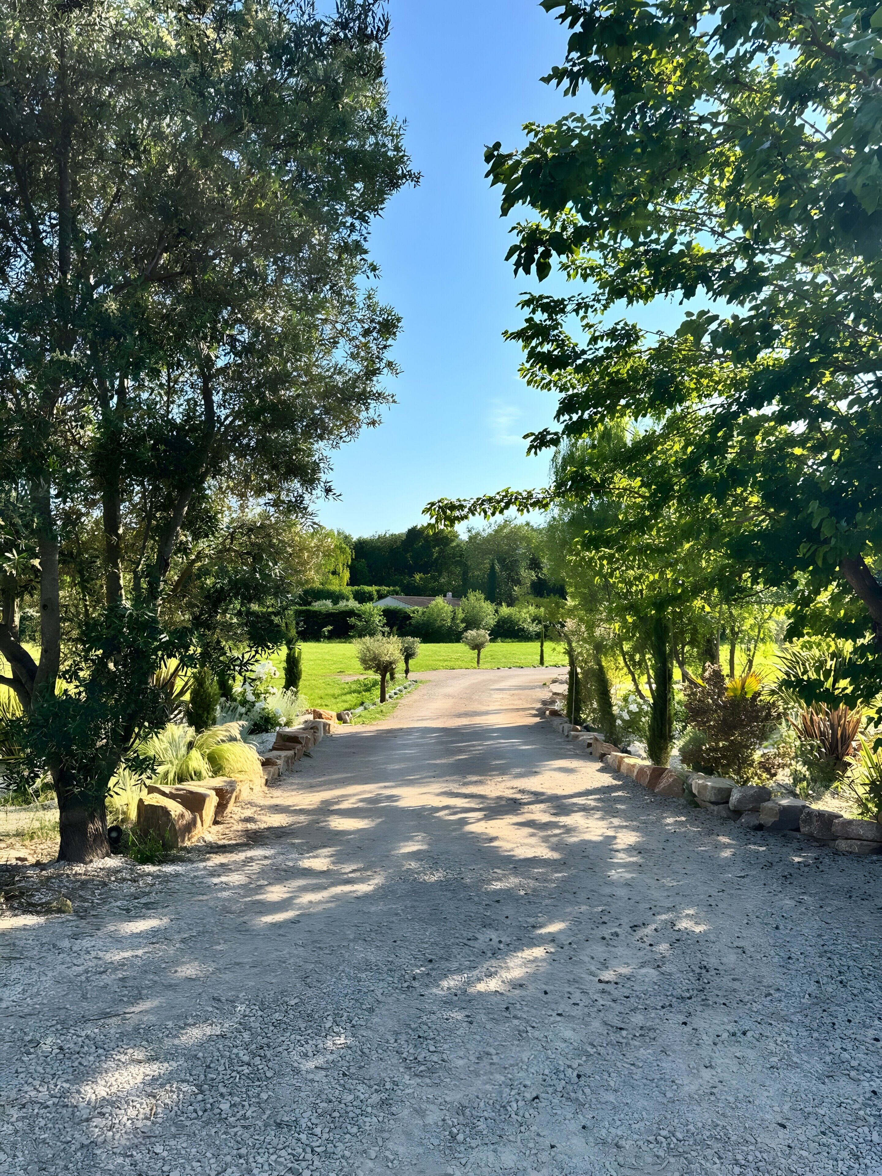Un chemin de gravier bordé d’arbres et d’arbustes, qui mène à un bâtiment situé dans un espace paysager.