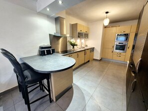 A compact kitchen with a built-in island, modern appliances, and a dining area with a chair. - Holiday Home 'Domaine Montserrat' with Mountain View, Private Pool and Wi-Fi (Le Luc)