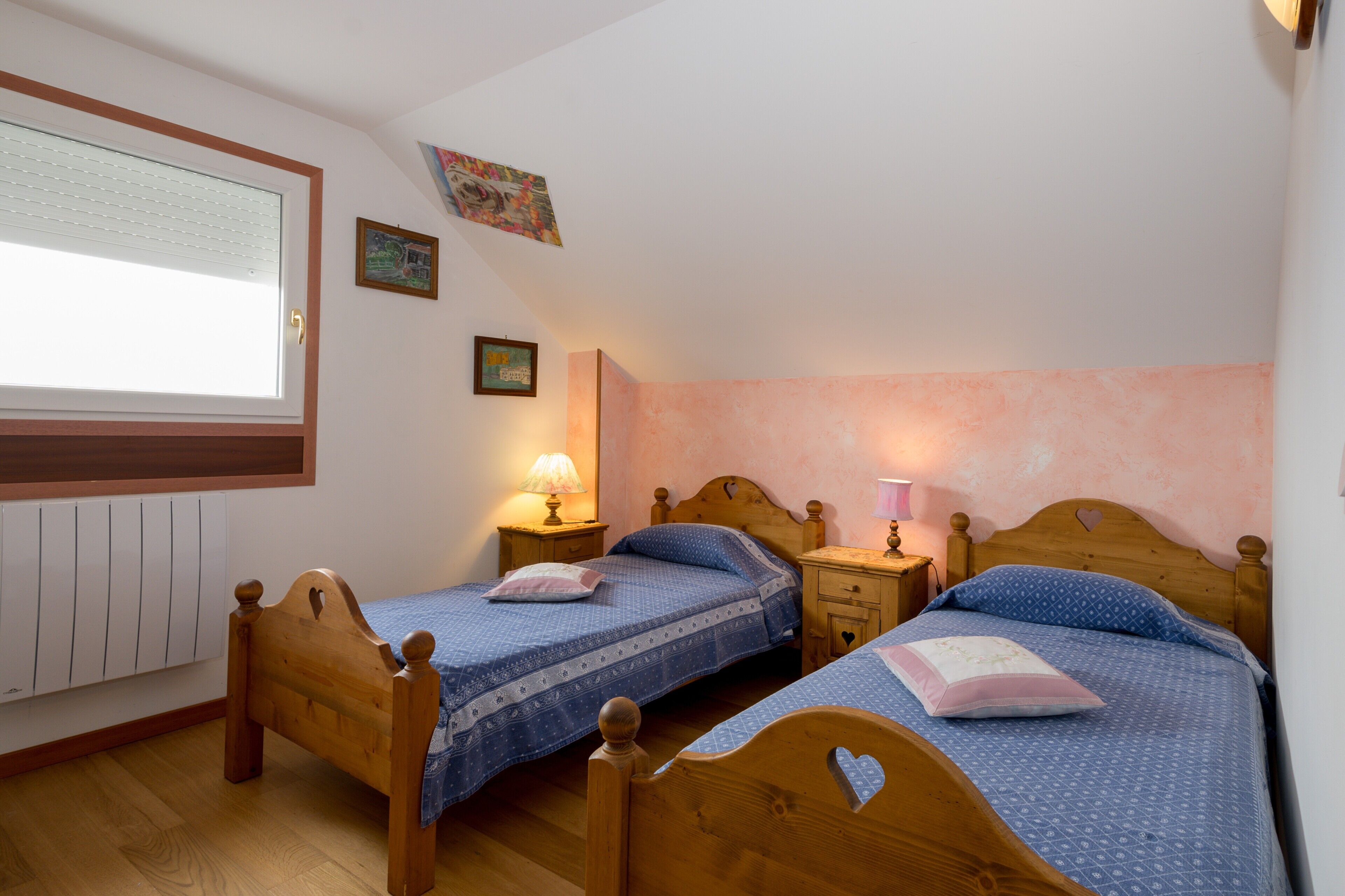 A bedroom with two wooden beds, a window with blinds, a radiator, and wall-mounted lamps.