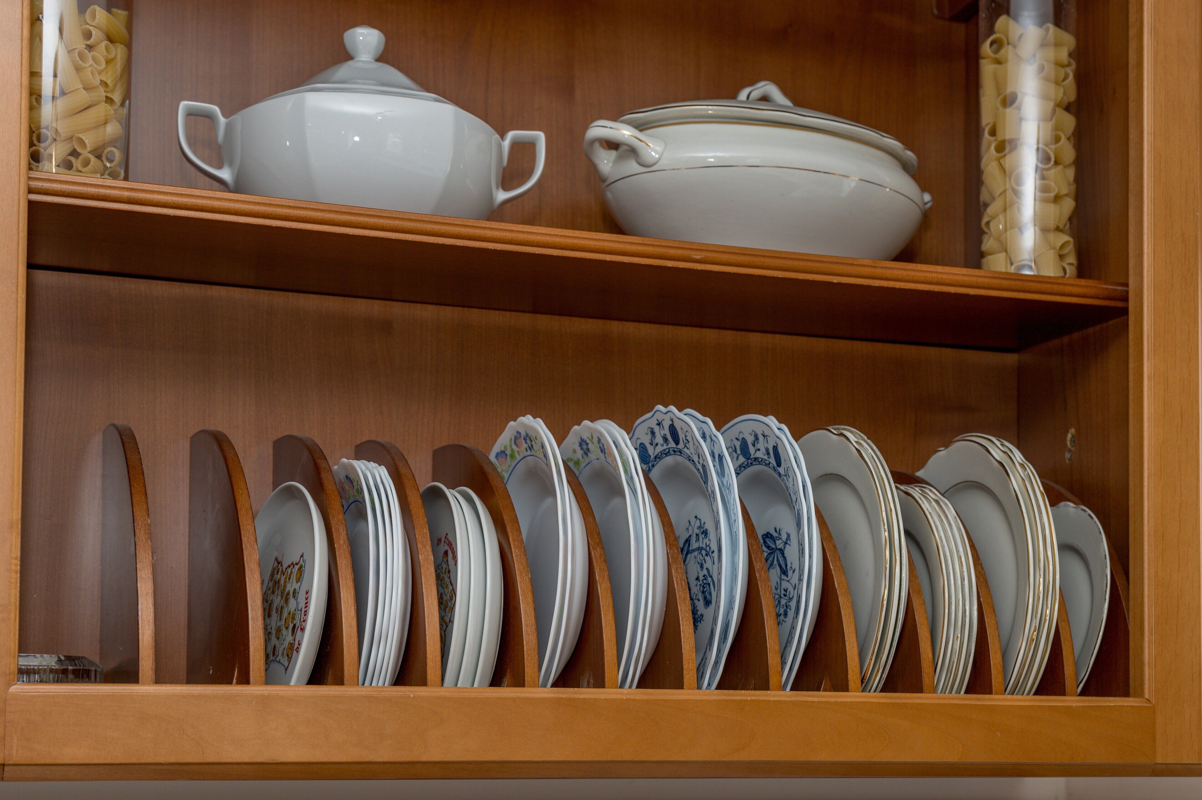 Una estantería de madera con utensilios de cocina de cerámica blanca, incluyendo una olla y un bol, y una fila de platos apilados con diseños florales.