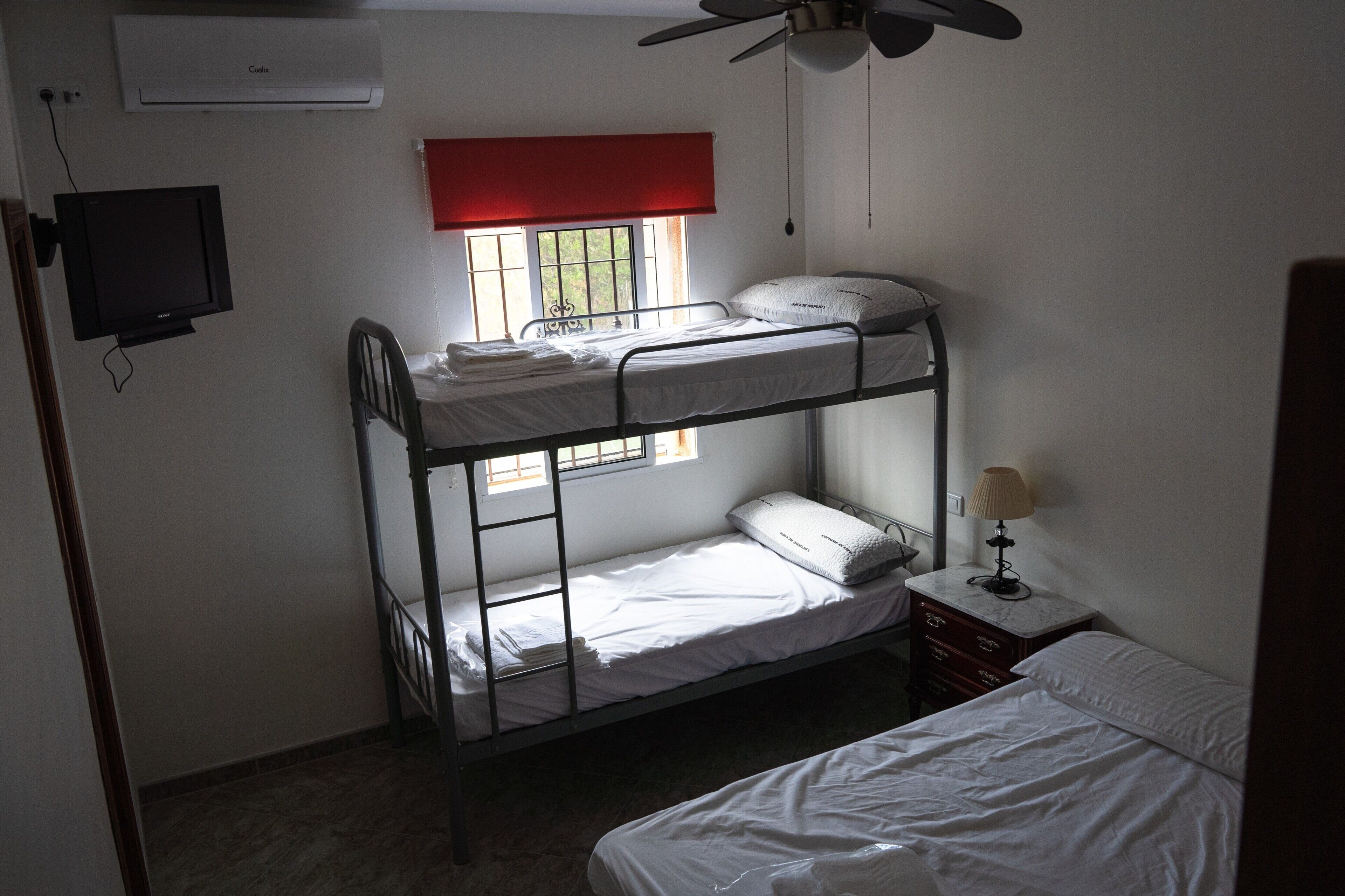 Habitación con literas, televisor, ventana con persiana roja y ventilador de techo.