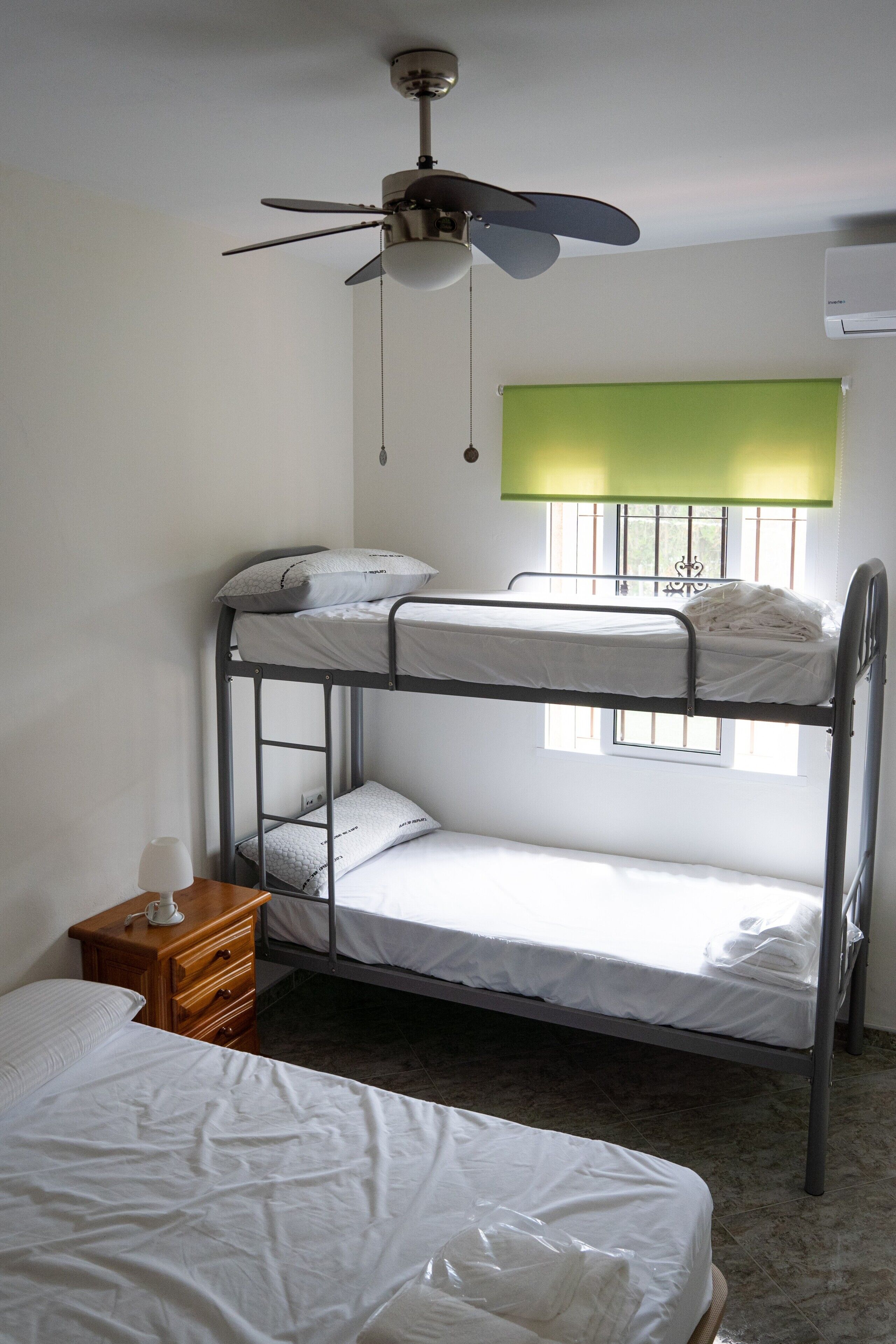 Habitación con literas, ventilador de techo, ventana con barrotes y mesita de noche con lámpara.
