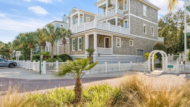 A multi-story building with a white porch and balcony, surrounded by palm trees and a white fence.