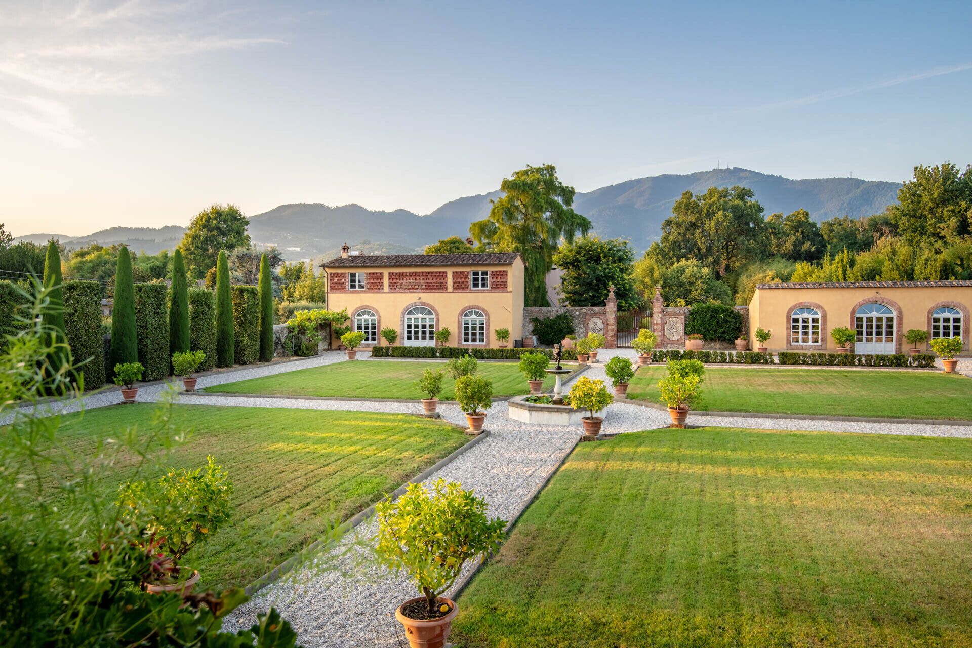 A well-maintained garden with a central pathway, a building with arched windows, and a mountainous backdrop.