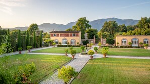 A well-maintained garden with a central pathway, a building with arched windows, and a mountainous backdrop. - Air Conditioned 10 Bedrooms Luxury Villa and Pool in Lucca (Capannori)