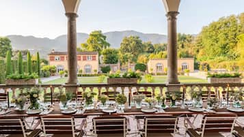 An outdoor dining area with tables set for a meal, overlooking a landscaped garden and distant buildings.