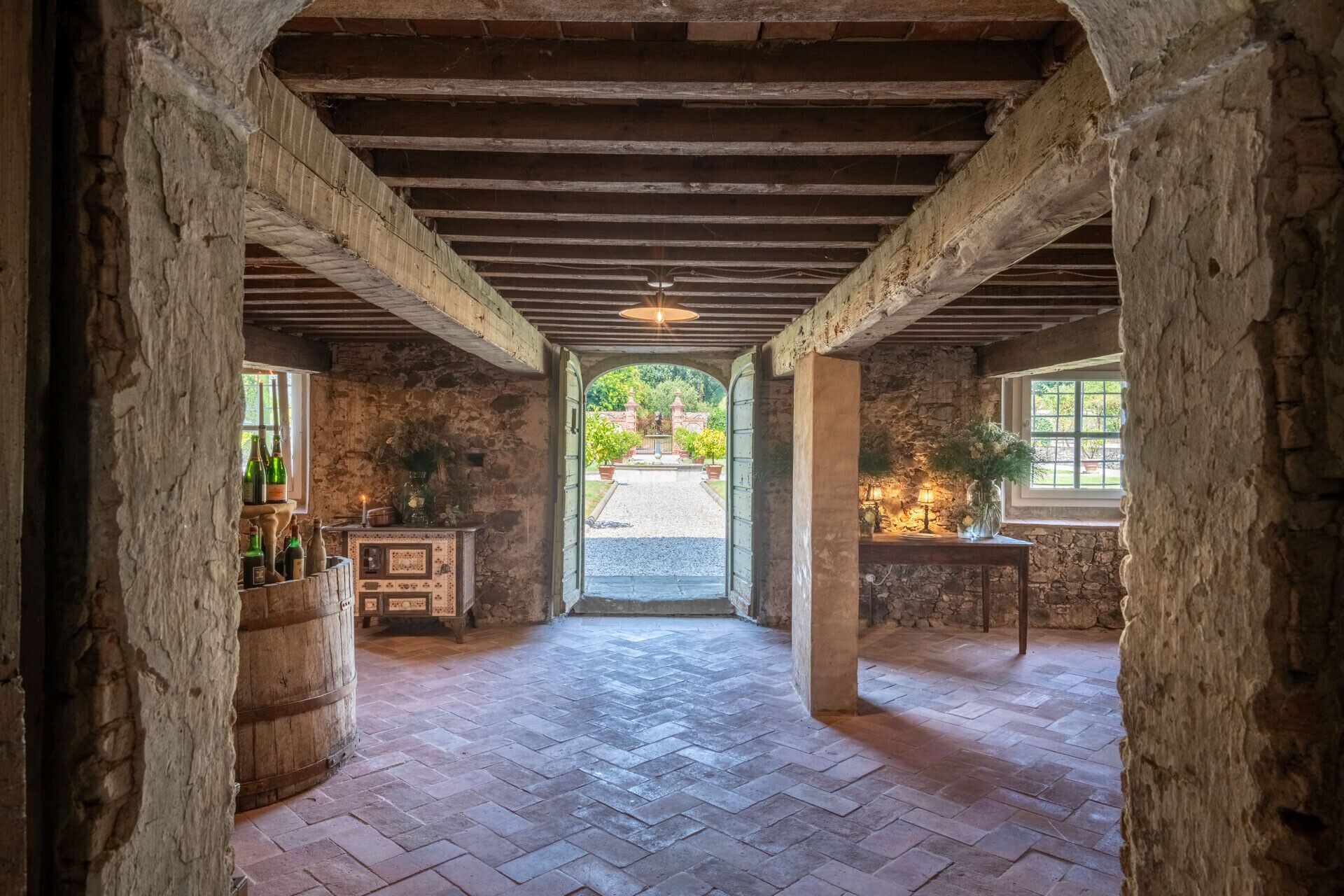 A rustic stone-walled room with a herringbone floor, wooden beams, and a view of a garden through an arched doorway.