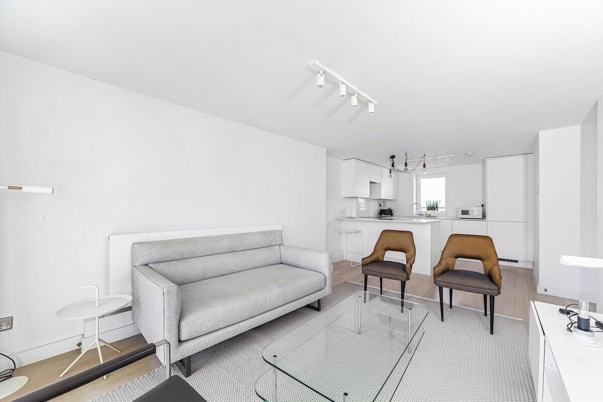 A modern living room with a grey sofa, two brown chairs, a glass coffee table, and a kitchen in the background.