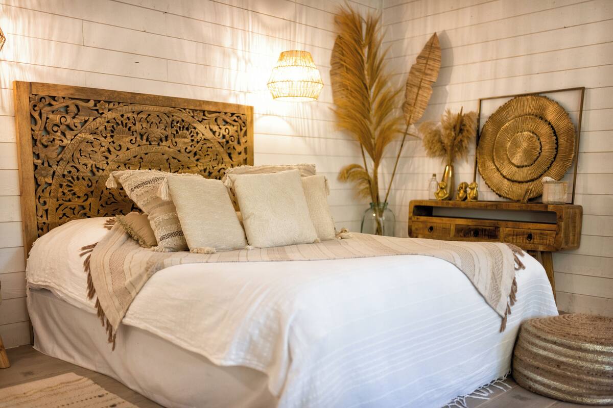 A bedroom with a wooden headboard, a large bed with multiple pillows, a bedside table with decorative items, and a wall-mounted lamp.