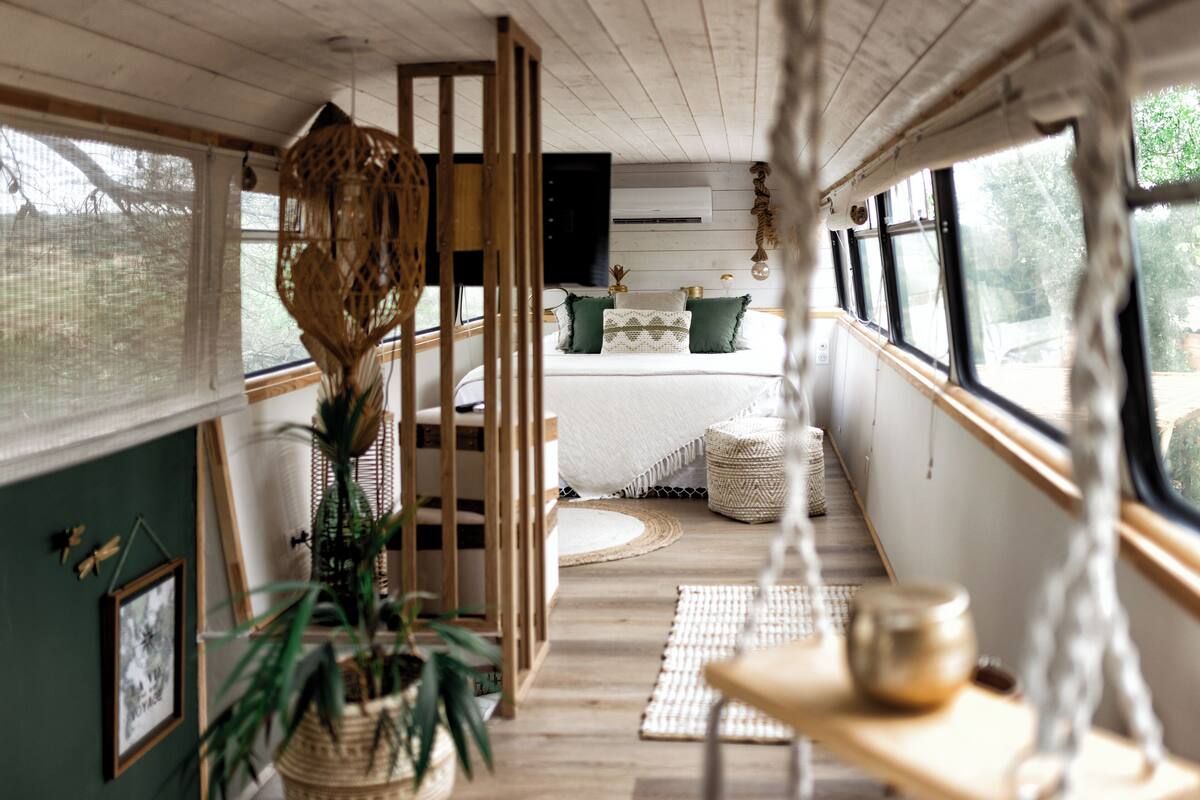 A narrow, wooden-paneled room with a bed, a hanging chair, and a window overlooking greenery.