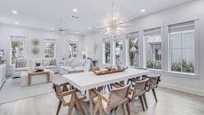 Un salón amplio con un sofá blanco, una mesa de comedor con sillas de madera y grandes ventanas con vistas a palmeras.