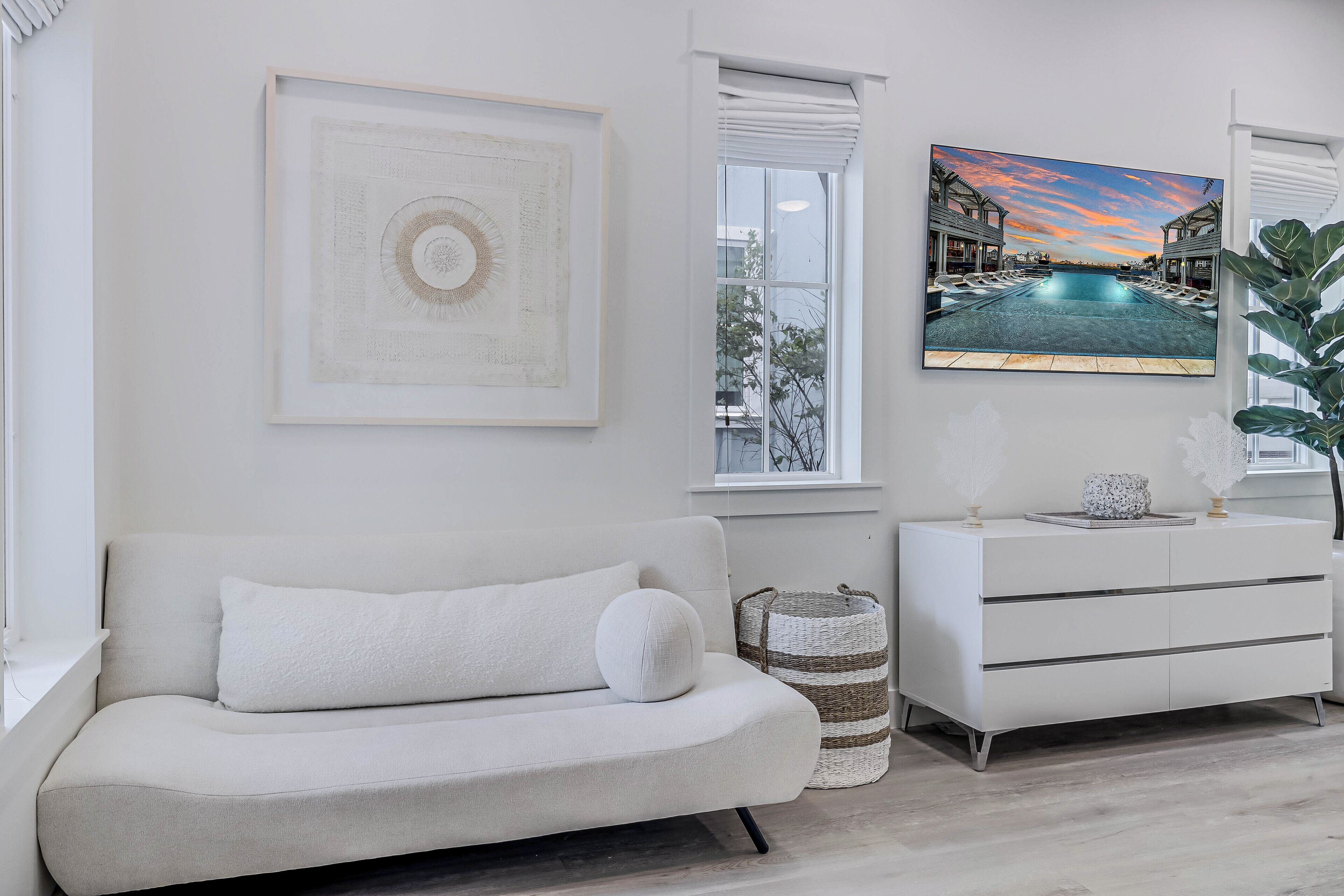 Uma sala de estar moderna com um sofá, uma cômoda branca, uma obra de arte abstrata emoldurada e uma grande janela com vista para uma piscina e edifícios.