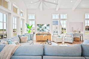 Ein großzügiges Wohnzimmer mit einer großen, grauen Eckcouch, einer Holzkommode und Blick auf Palmen durch große Fenster.