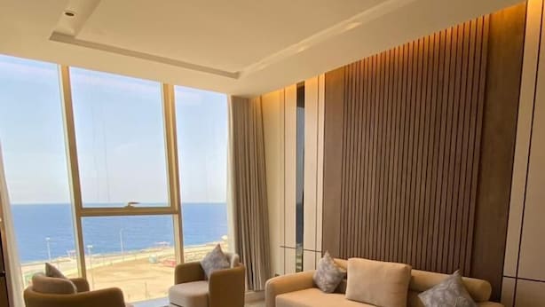 Un salon moderne avec un canapé beige, deux fauteuils et une grande fenêtre donnant sur une plage.