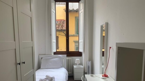 Une chambre d’hôtel étroite, avec un lit, un bureau, un radiateur et une fenêtre donnant sur les bâtiments environnants.