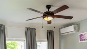 Une chambre à coucher avec un lit, des rideaux gris, un ventilateur de plafond, une table de chevet avec une lampe et une fenêtre donnant sur un paysage verdoyant.