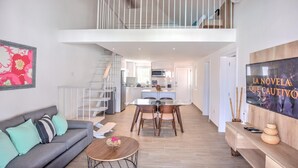 A spacious living area with a grey sofa, a dining table with chairs, a staircase leading to an upper level, and a kitchen area. - TesCosy Beachfront 2 BDR Apartment. STM G406 (BAVARO)