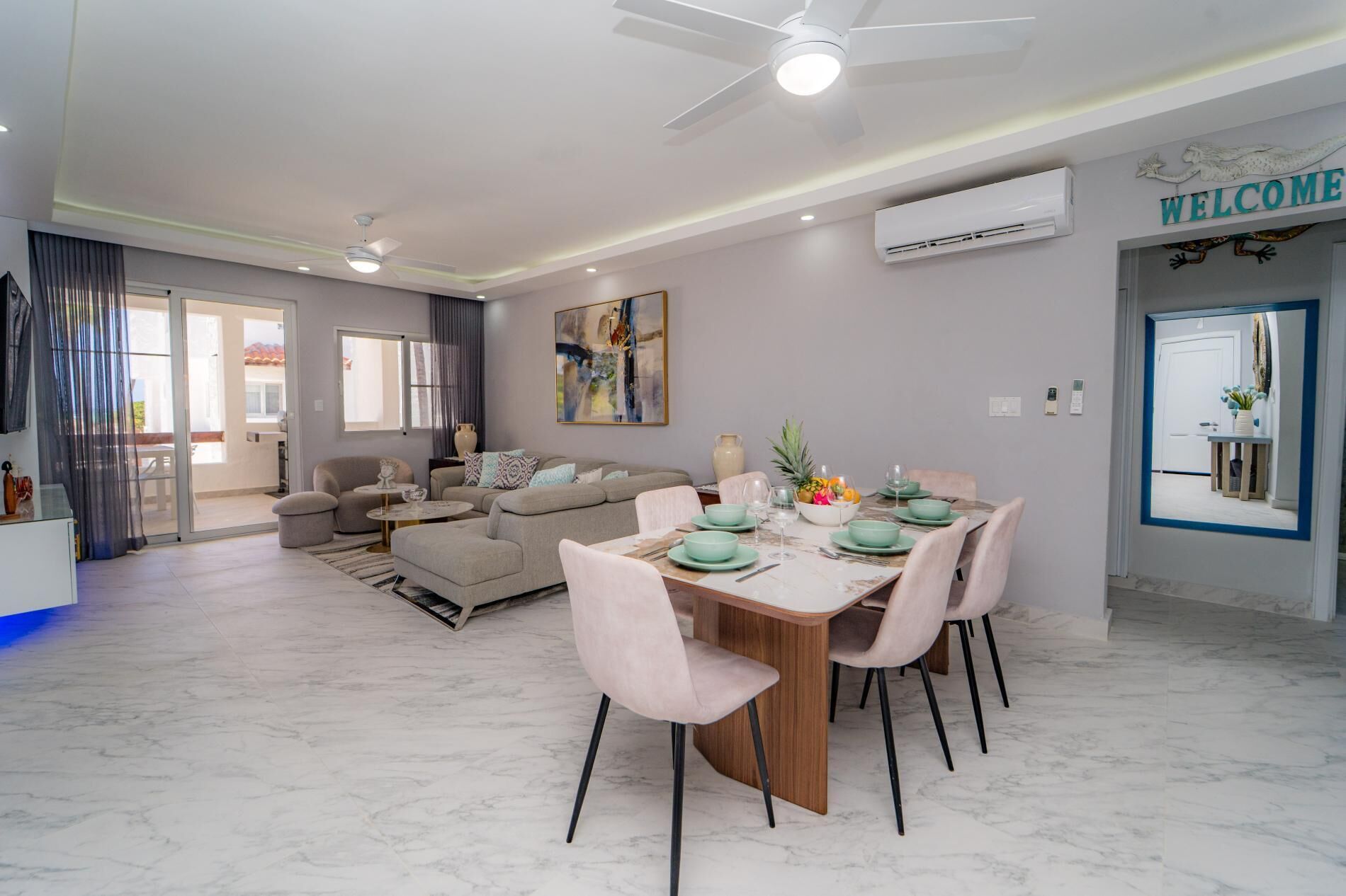 A modern living room with a dining area, a ceiling fan, and a welcome sign.