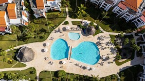 Vue aérienne d’un complexe hôtelier avec deux grandes piscines, des palmiers et les bâtiments du complexe.