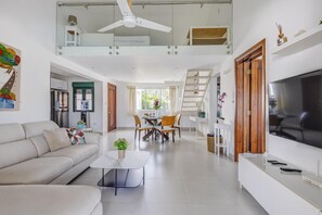 Een ruime woonkamer met een bank, salontafel, eettafel en stoelen. Een televisie is aan de muur bevestigd, en er is een trap die naar een hoger niveau leidt.