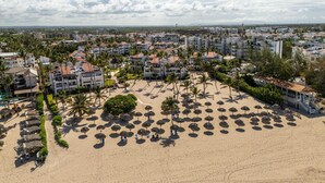 A coastal resort with multiple buildings, a sandy beach with thatched umbrellas, and palm trees. - Room Mare F-302  Share  Save (BAVARO)
