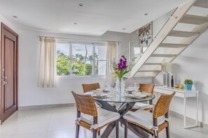 A dining area with a glass table, wicker chairs, a vase with flowers, a staircase, and a view of palm trees.