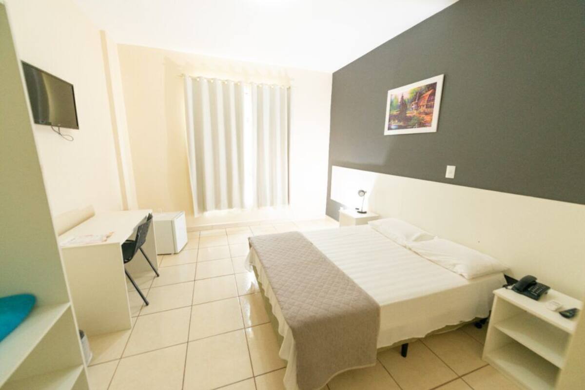 A hotel room with a bed, a desk, a chair, a TV, a bedside table, and a window with curtains.
