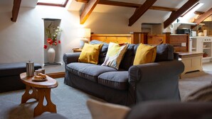 Una sala de estar acogedora con un sofá gris, vigas de madera, tragaluces, un jarrón con flores y una pequeña mesa de madera con una tetera y pasteles.