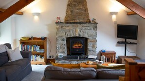 Un salón acogedor con chimenea de piedra, vigas de madera, estantería, sofá y televisor.