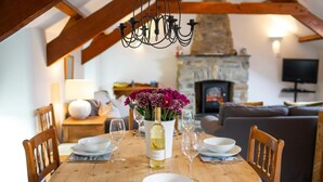Mesa de comedor preparada para cuatro personas, con platos, tazones y copas de vino blancos, una botella de vino y un florero con flores moradas. La habitación tiene sillas de madera, una chimenea de piedra y un televisor en la pared.