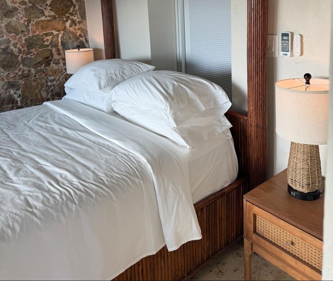 A neatly made bed with white linens, a wooden headboard, a bedside lamp, and a stone wall.