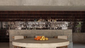 Ein runder Betontisch mit einer Obstschale in der Mitte, umgeben von Stühlen. Hinter dem Tisch ist eine mit Flaschen und einem Metallgitter versehene Wand zu sehen.